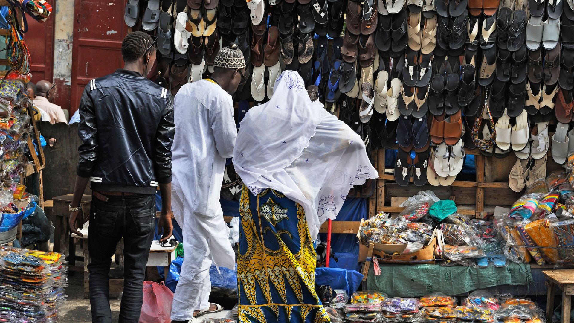 Two men are inspecting shoes at a vibrant market, surrounded by other shoppers.