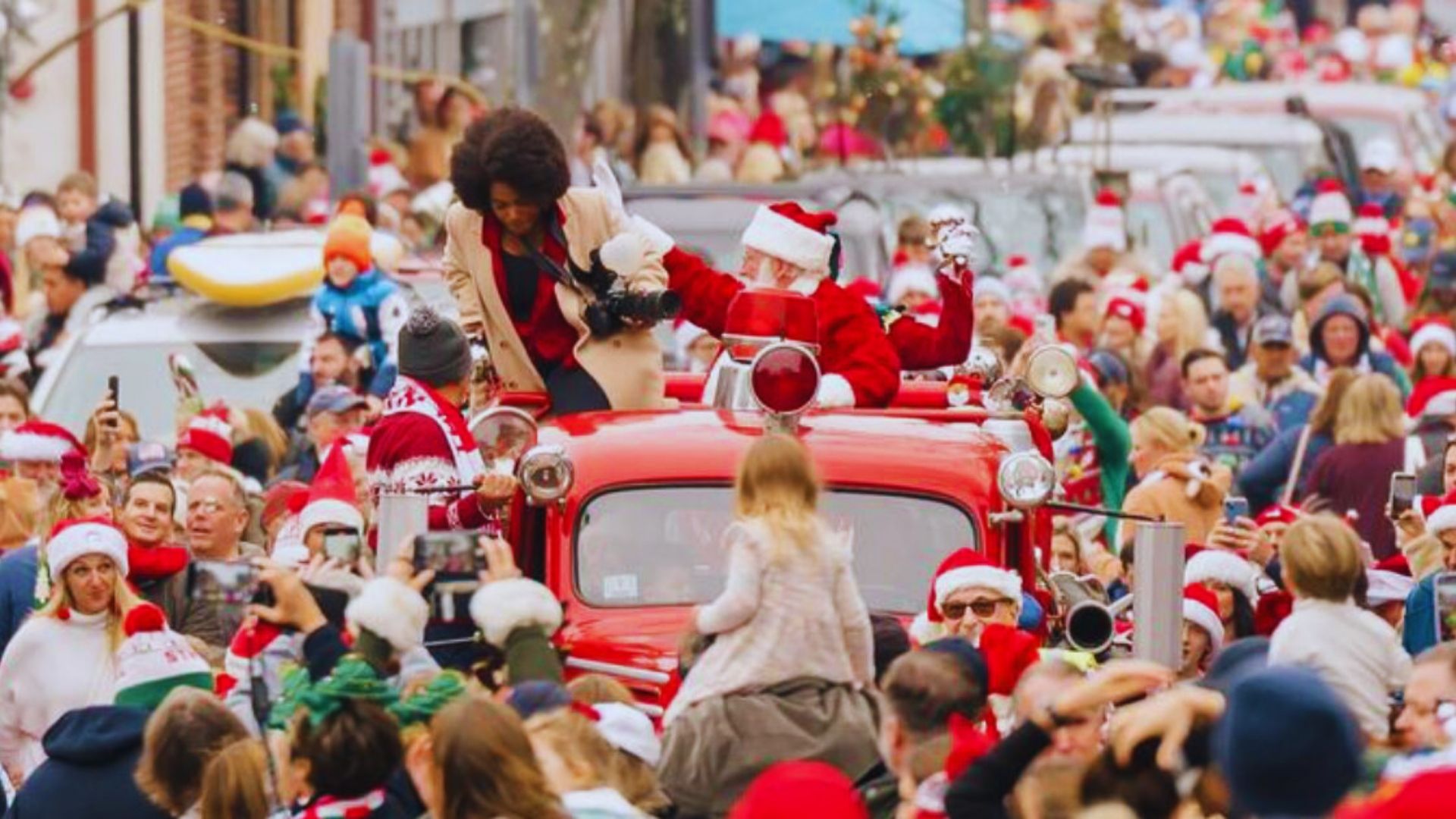 A lively crowd wearing Santa hats and suits gathers on a festive street, celebrating the holiday spirit.