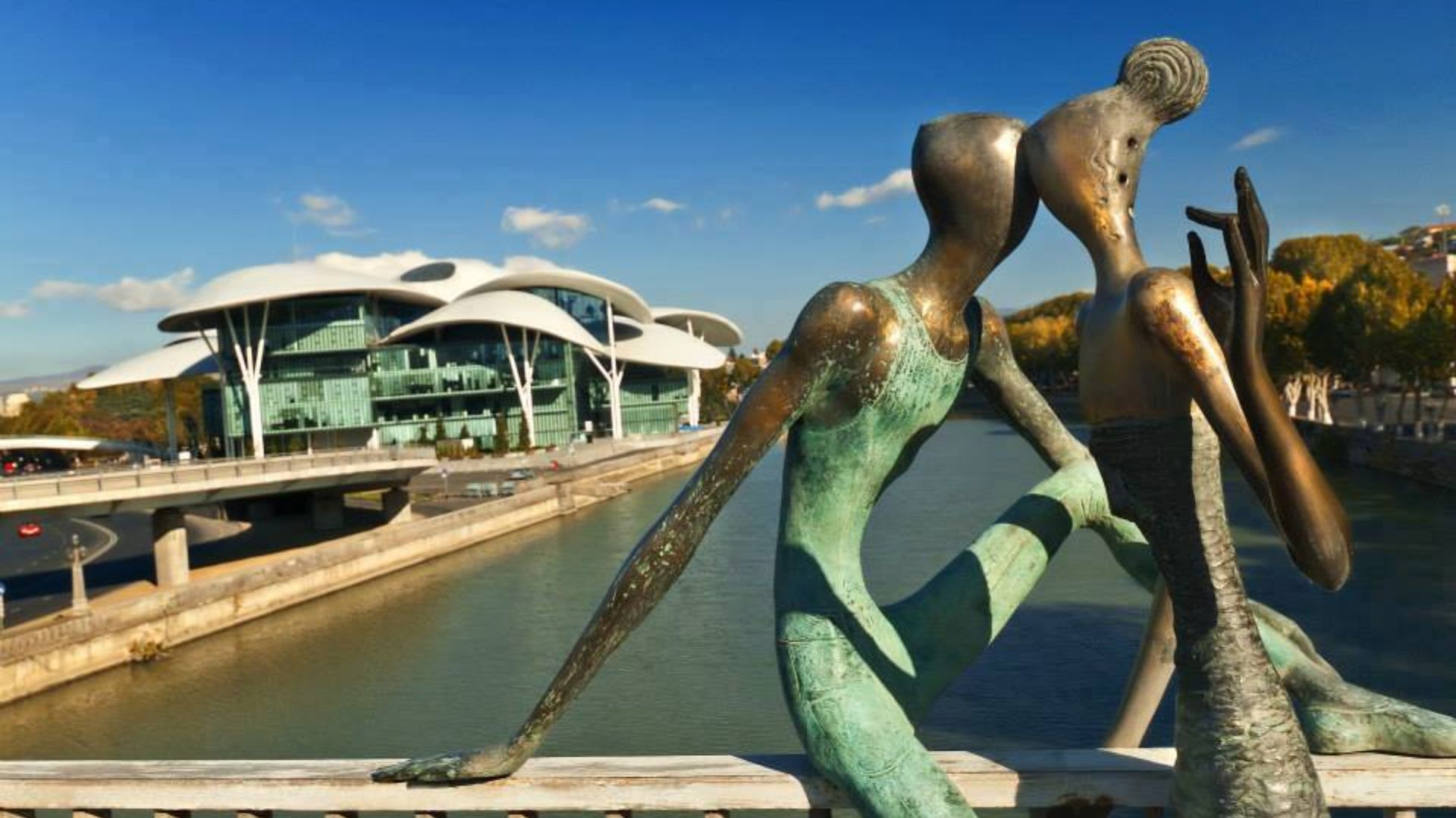 Two statues of people seated on a bridge overlooking a river, capturing a serene moment in a scenic outdoor setting.