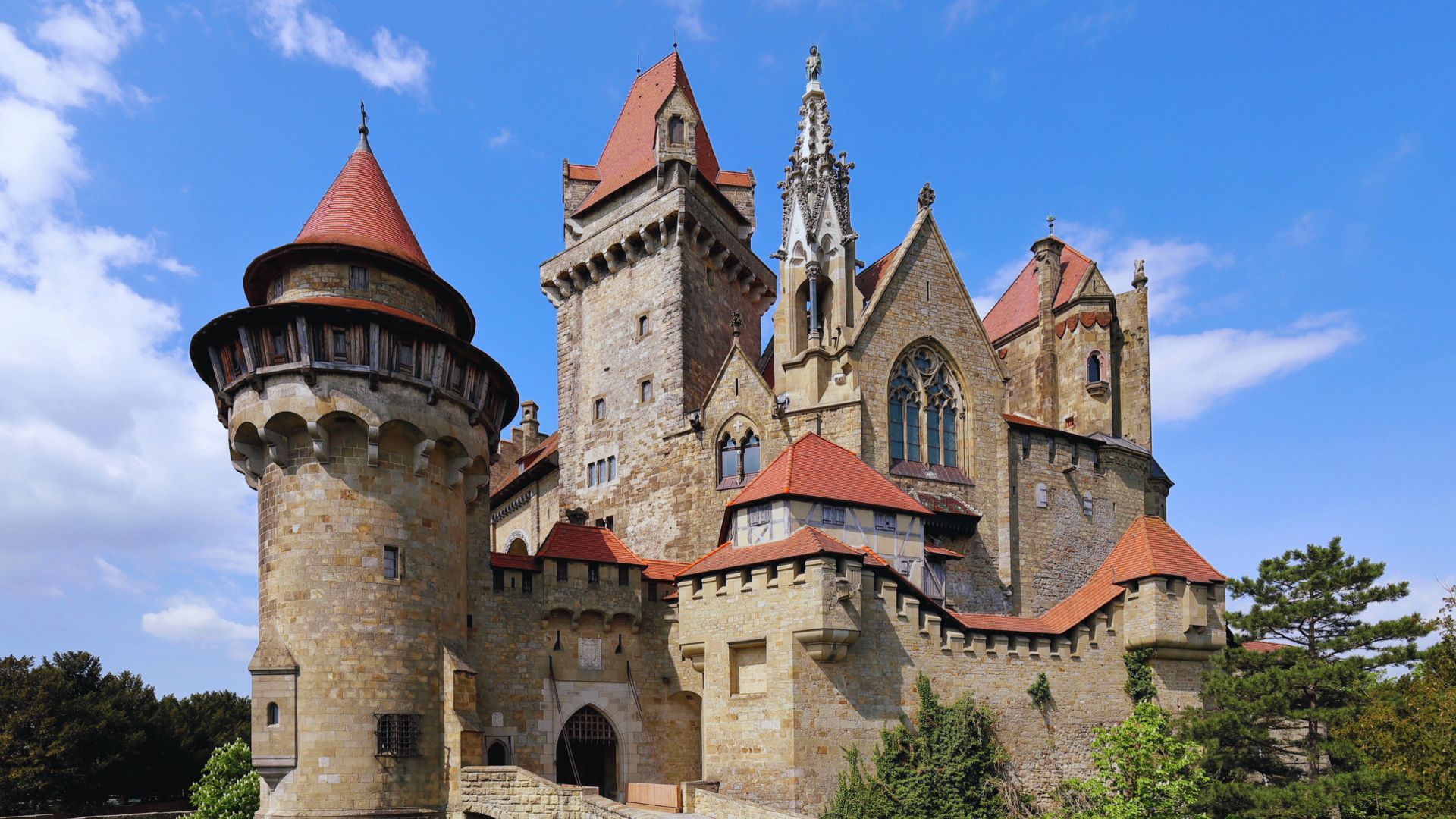 Medieval castle with stone towers and red-tiled roofs against a blue sky. The architecture is ornate, featuring pointed arches and intricate details.