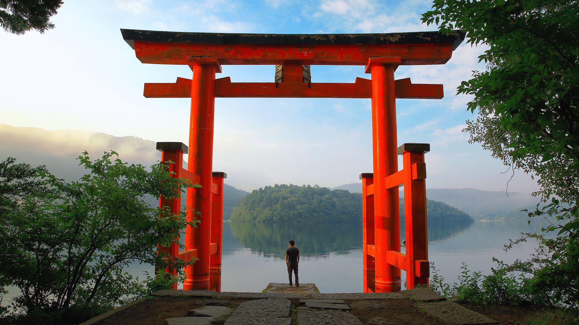 A person stands before a large red torii gate overlooking a serene lake and misty hills. The scene is tranquil, with lush greenery framing the view.