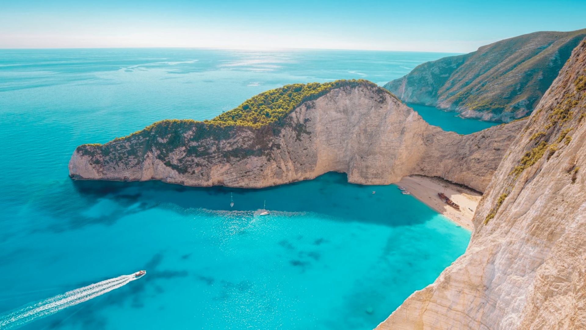 Aerial view of a serene cove with turquoise waters surrounded by steep, rocky cliffs and lush greenery. A few boats leave gentle wakes in the calm sea.