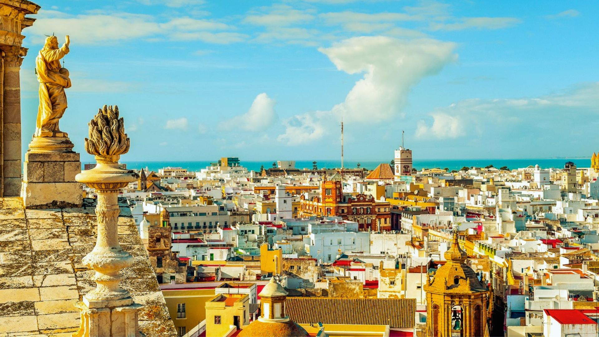 Rooftop view of a vibrant cityscape against a bright, partly cloudy sky. Ornate statues and architectural details are in the foreground, conveying historical charm.