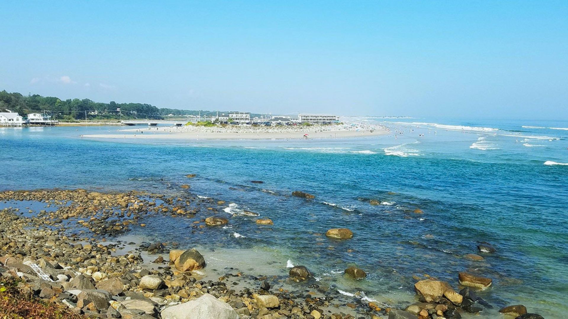 Coastal scene with rocky shoreline in foreground, waves gently lapping at the beach, and blue sky overhead. Buildings and trees line the distant shore.