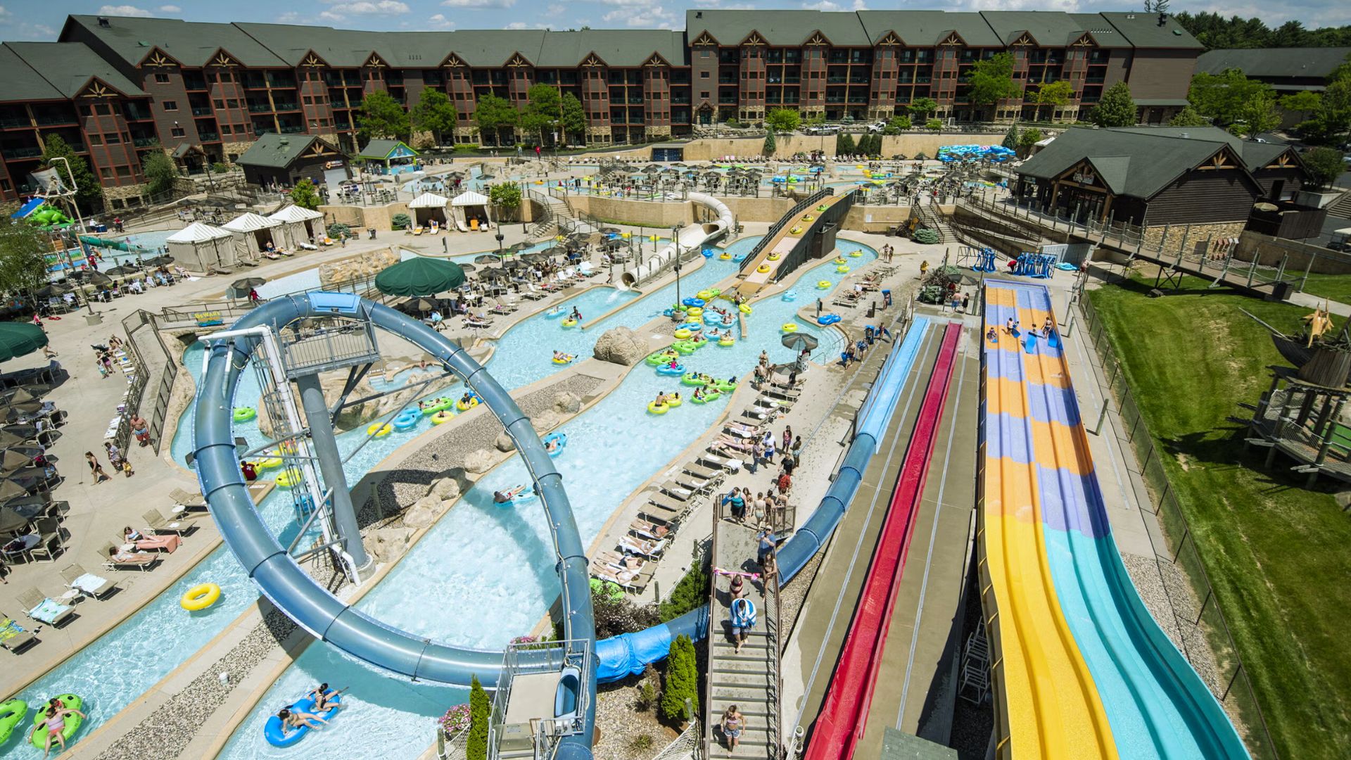 Aerial view of a vibrant water park with colorful slides and a lazy river, surrounded by loungers and a lodge-style building, creating a lively and fun atmosphere.