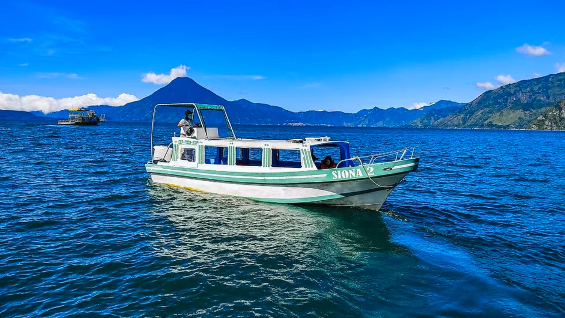 A white and green boat named "Siona 2" floats on a vast blue lake, with a distant volcano and lush mountains under a bright blue sky. Calm and scenic.