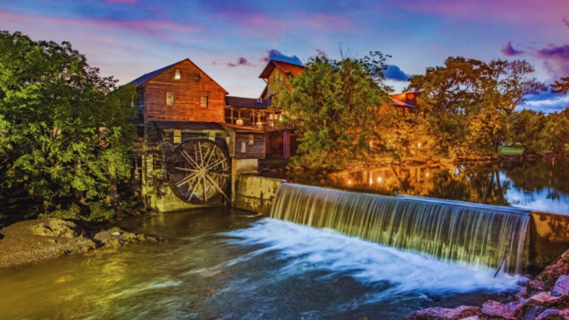 A rustic watermill with a large wheel stands beside a small waterfall at sunset, surrounded by lush trees and reflecting a serene, colorful sky.