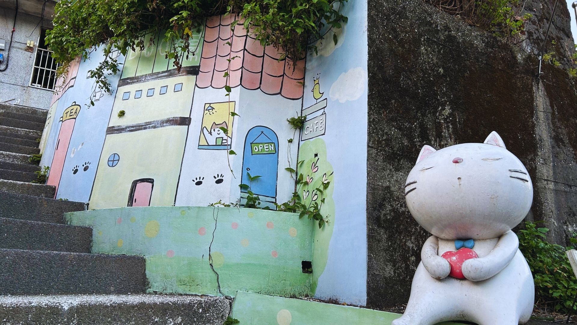 Whimsical mural on a wall with colorful houses, plant details, and paw prints. A large, cheerful white cat statue holds a red heart, adding playfulness.