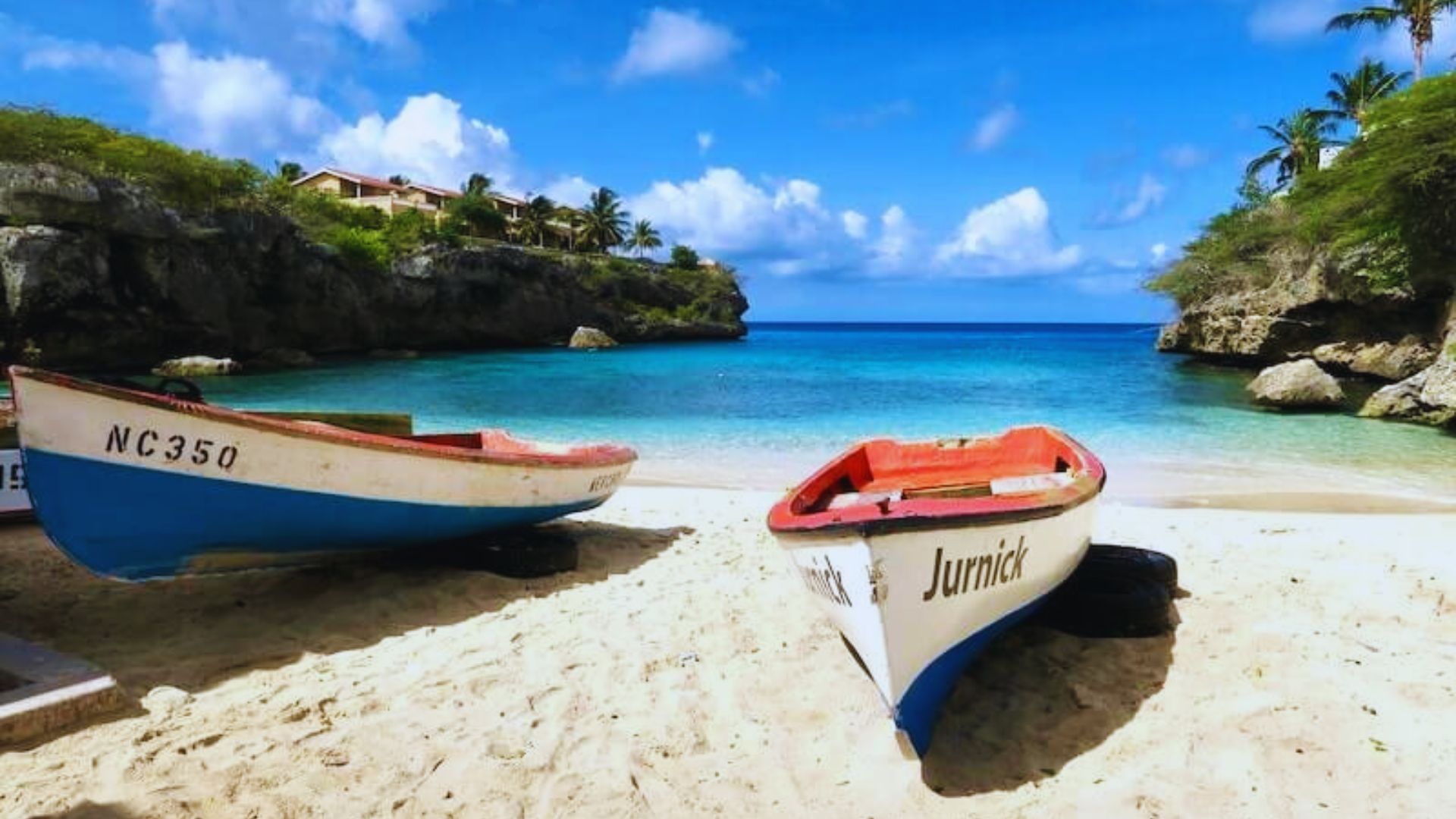Two colorful boats rest on a sandy beach next to clear turquoise waters under a bright blue sky. Lush greenery and rocky cliffs surround the scene, creating a tranquil and inviting tropical atmosphere.
