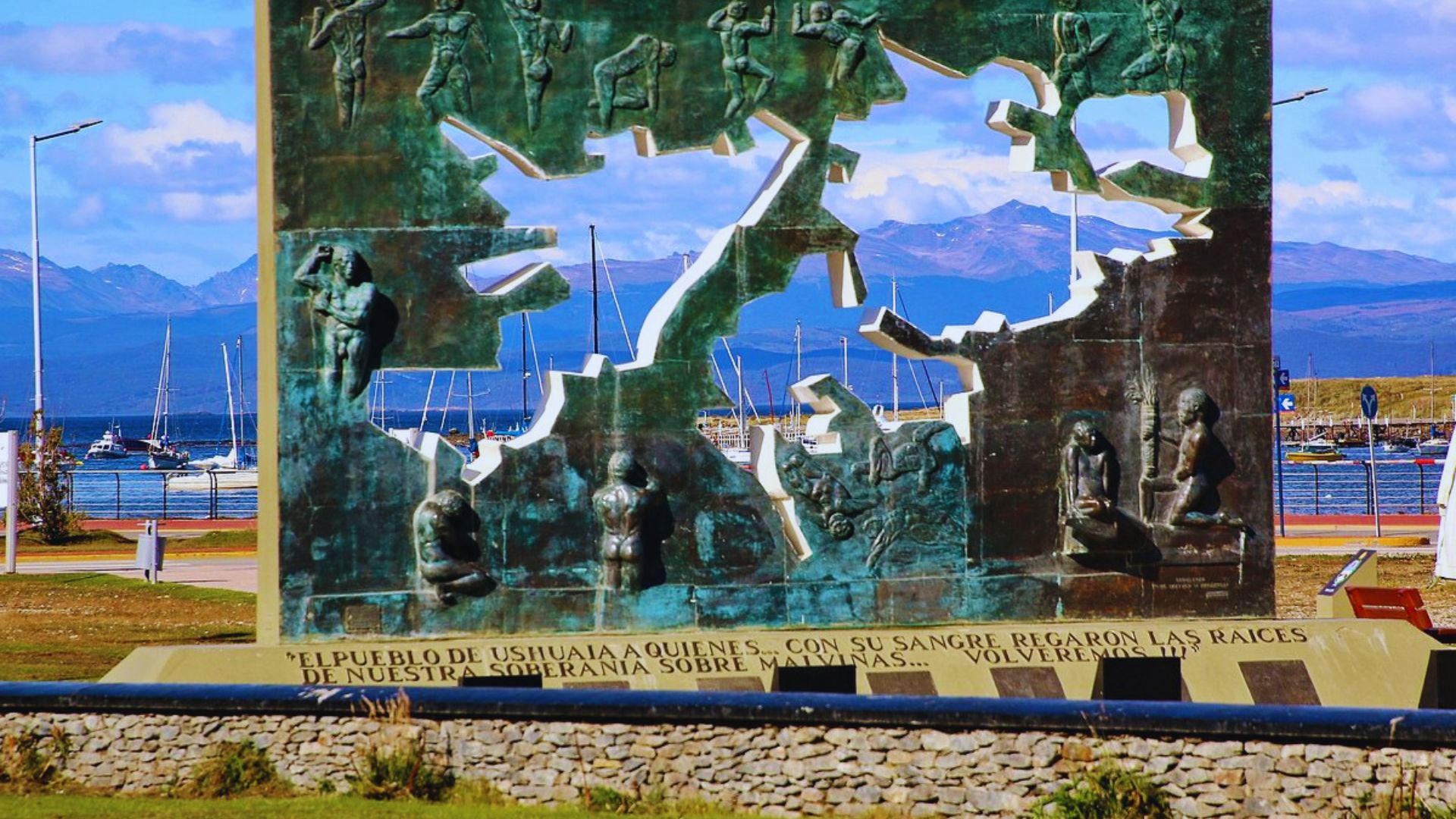 A bronze monument with figures and cutouts symbolizes Ushuaia's maritime history. Behind it, boats, mountains, and a clear blue sky set a peaceful tone.