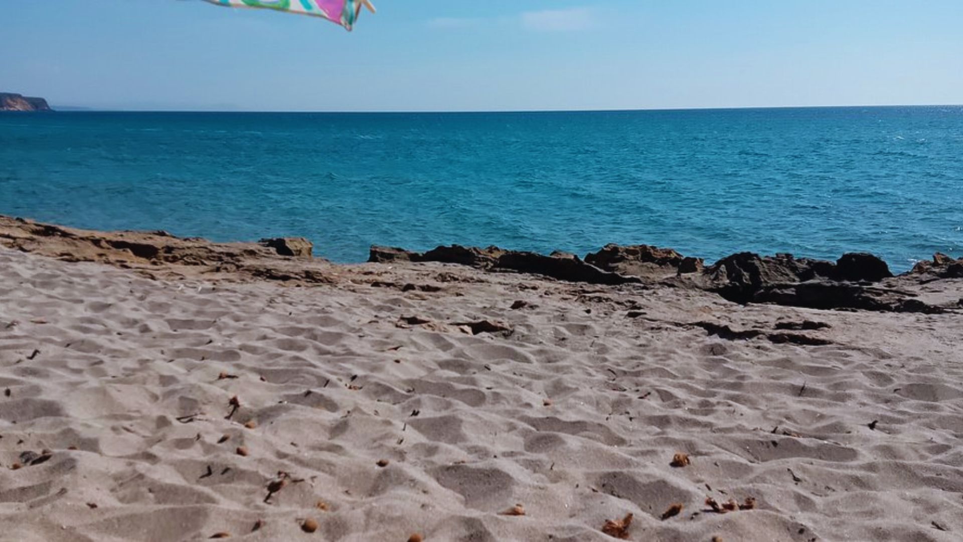 Sandy beach with turquoise ocean, clear blue sky, and rocks lining the shore. Vibrant towel waves from the top left, evoking a serene summer day.