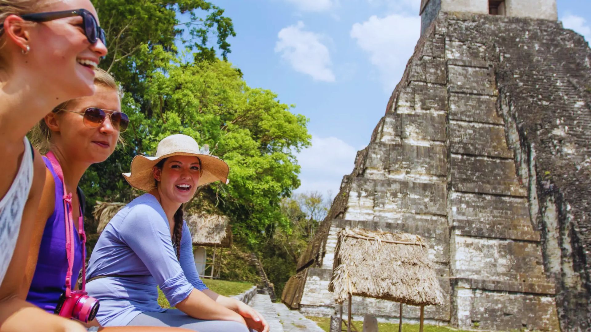 Three women sit on a bench, smiling, with a large pyramid visible in the background under a clear blue sky.