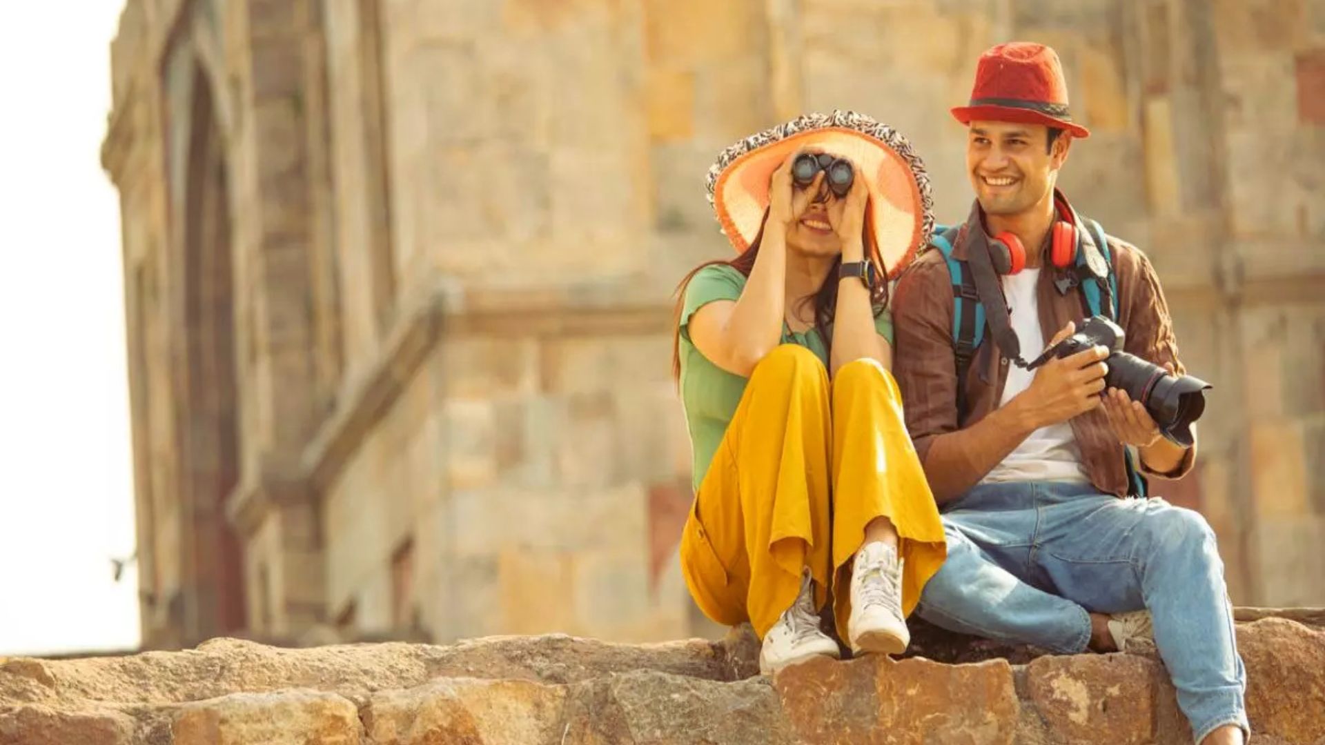 A couple sits on a wall, smiling and holding a camera, enjoying a moment together outdoors.