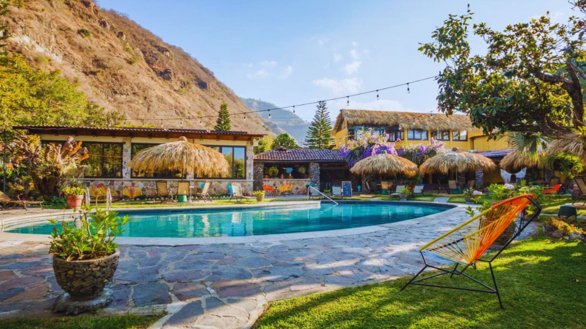 A serene mountain scene featuring a pool beside a thatched-roof house surrounded by lush greenery.