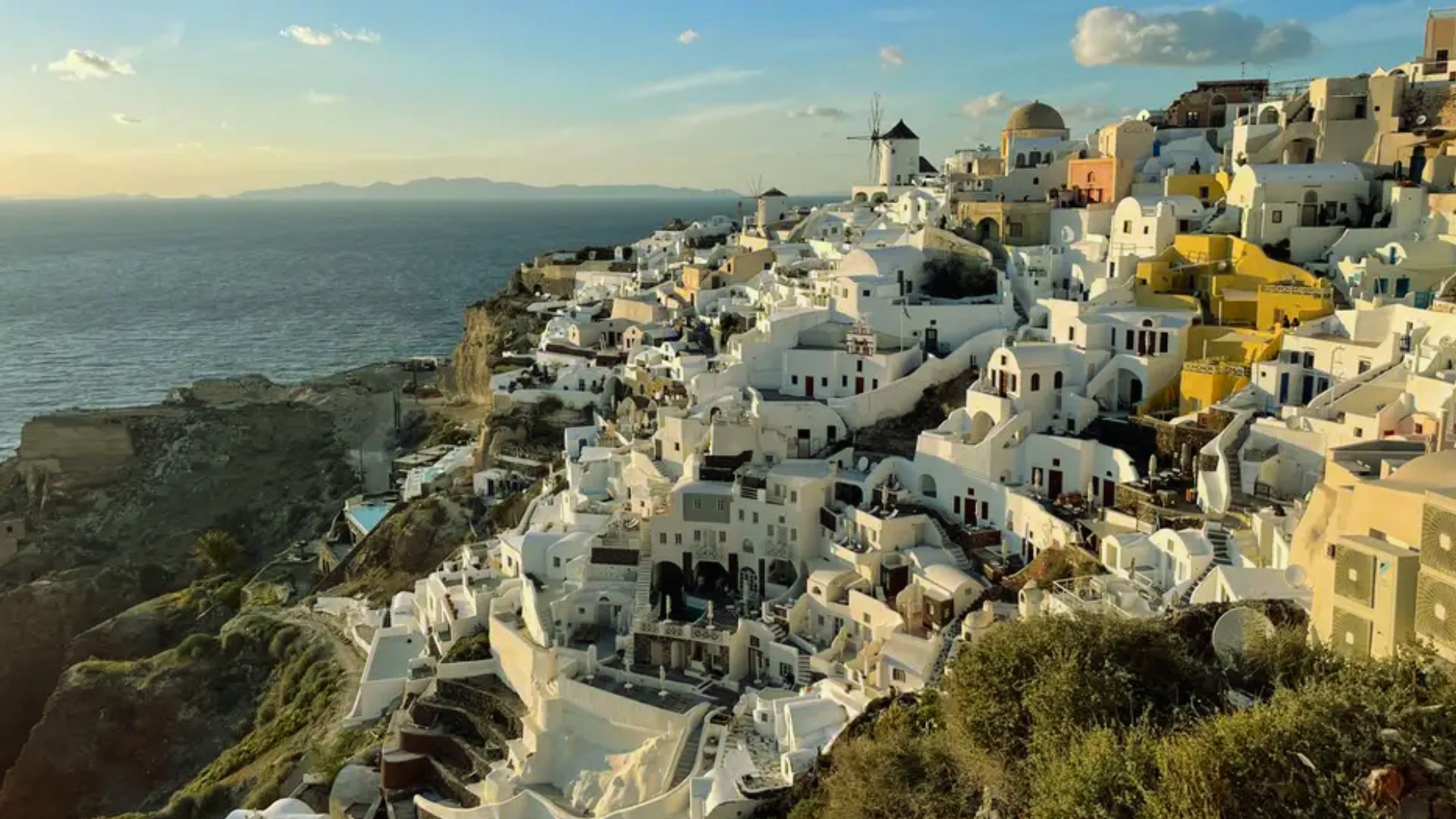 Scenic view of Santorini, Greece, with whitewashed buildings and blue domes cascading down a hillside at sunset. The Aegean Sea sparkles in the background.