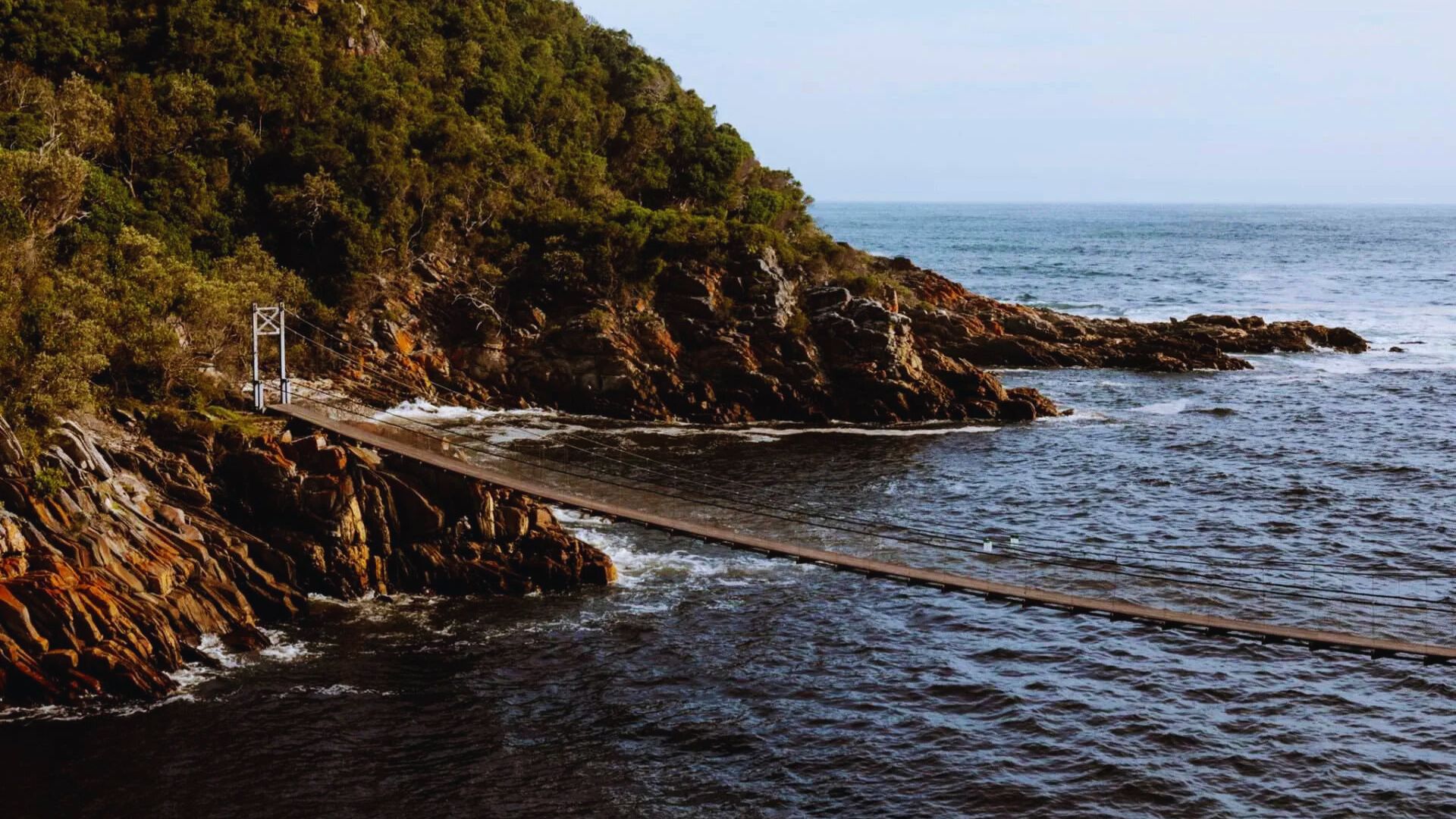 A rope bridge spans over the ocean, connecting the shore to a distant point, with waves gently lapping below.