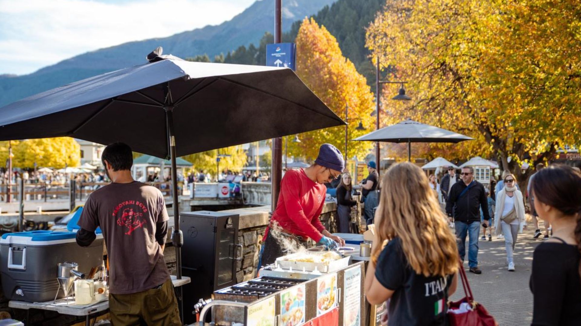A group of people gathers around a food stand with a colorful umbrella, enjoying the outdoor atmosphere.