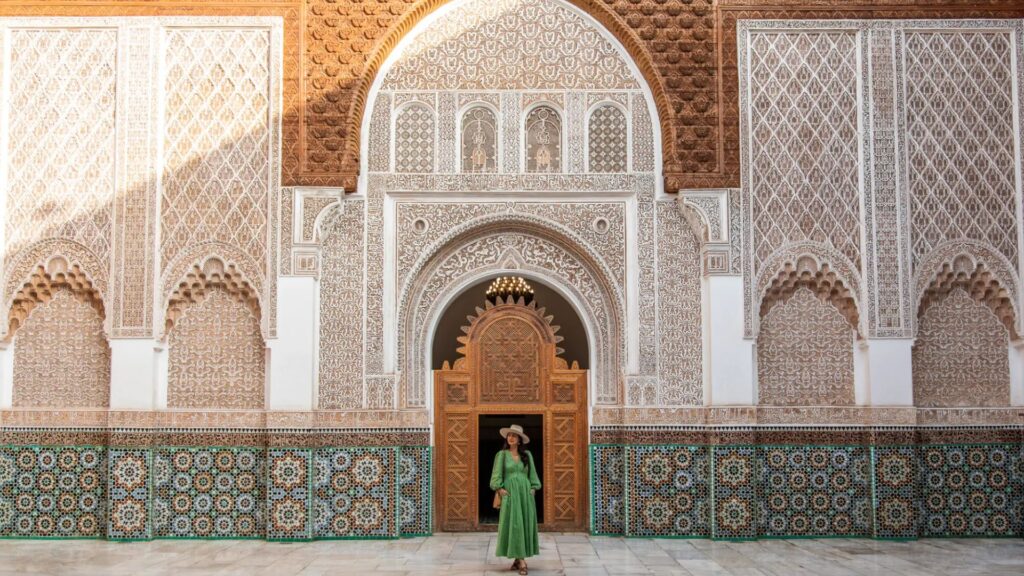 A woman in a green dress stands gracefully in front of an ornate doorway, exuding elegance and poise.