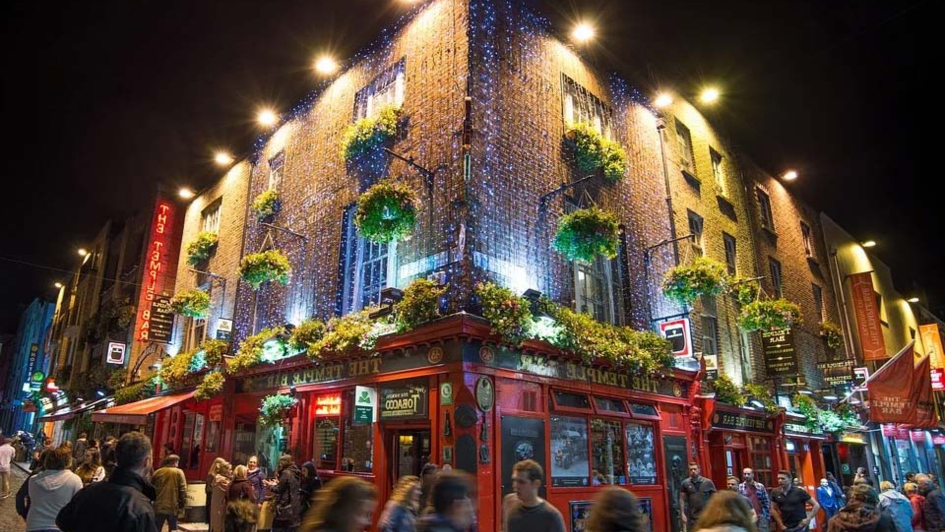 Nighttime scene of people walking along a street, with a pub illuminated in the background.
