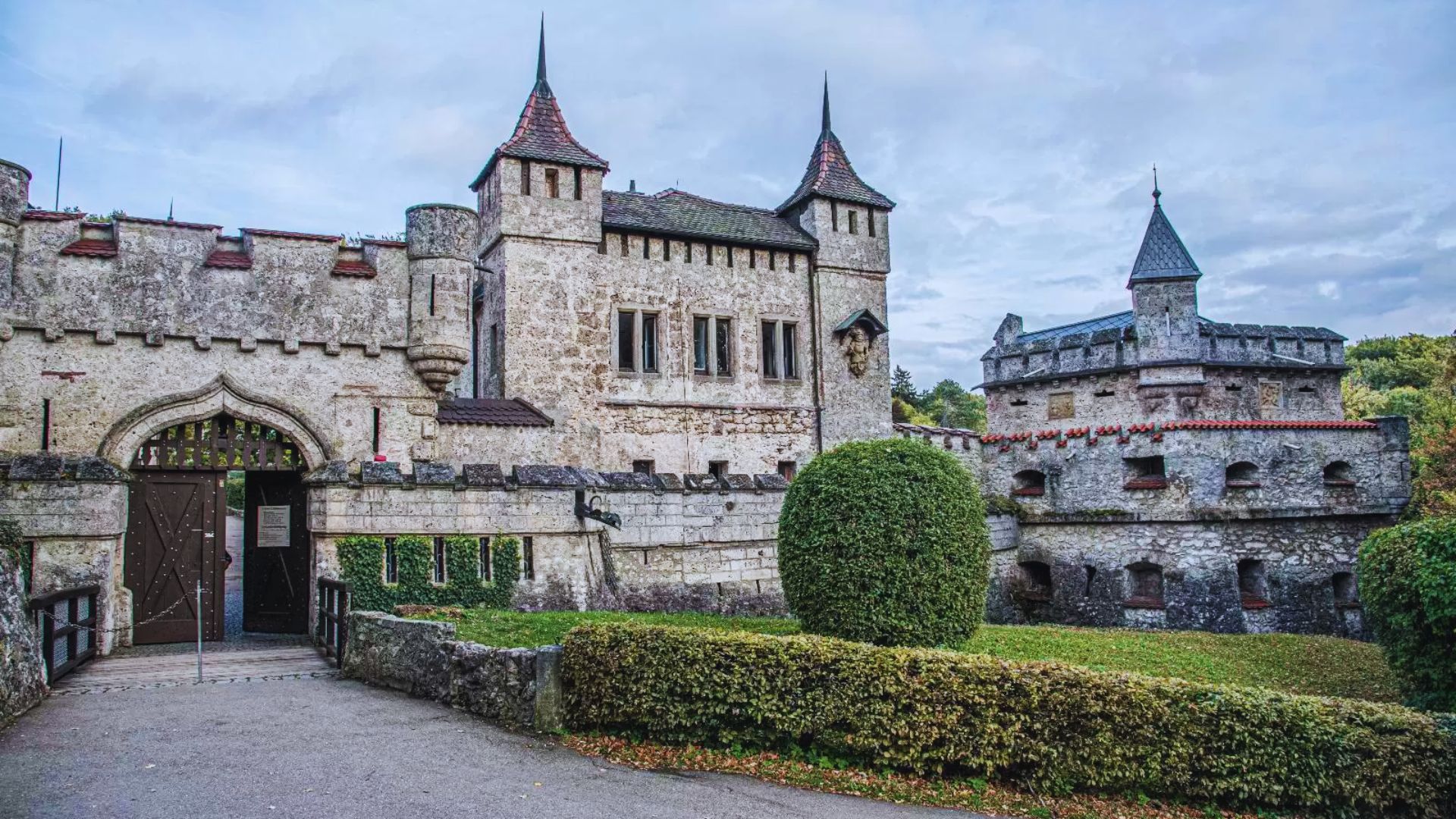 A historic stone castle with tall, pointed towers under a cloudy sky. A neatly trimmed hedge lines the front, adding greenery and contrast. Majestic and serene atmosphere.