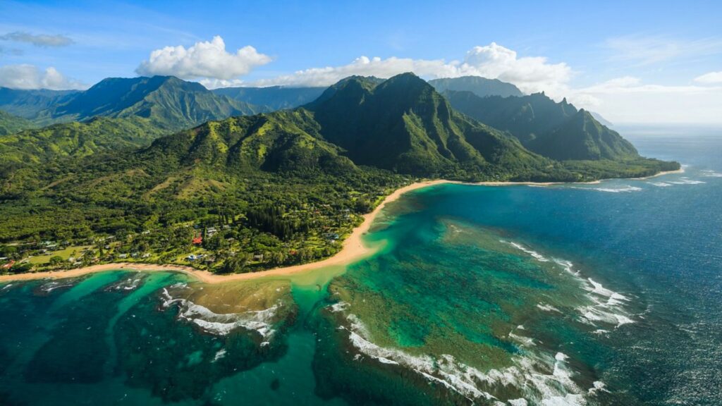 17 Things to Do in Hanalei: Top Attractions & Activities Aerial view of a lush coastline with turquoise waters and sandy beaches, backed by lush green mountains under a clear blue sky. The scene is serene and vibrant.