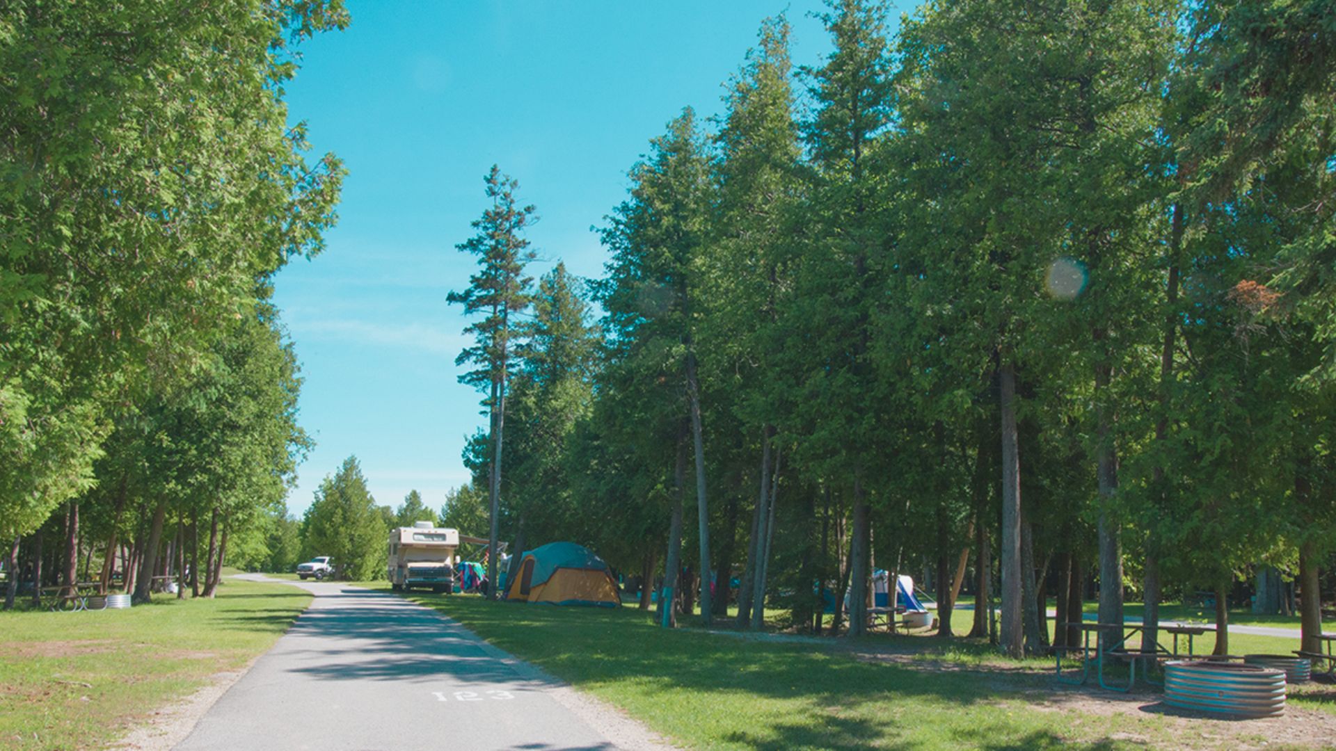 A sunny campsite with a paved road leading into the distance. Tents and trees line the right, with a parked RV visible on the left, evoking a sense of calm.