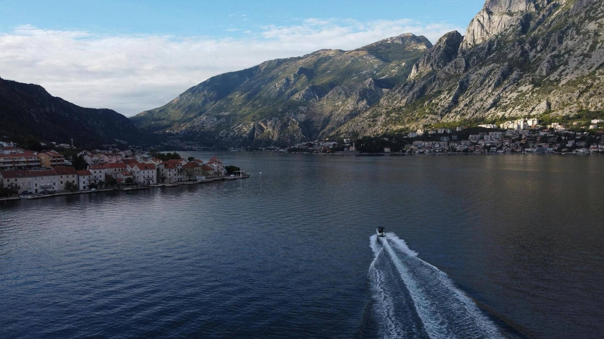 Aerial view of a serene bay surrounded by rugged mountains under a partly cloudy sky. A small boat leaves a trail on the calm water near a coastal village.