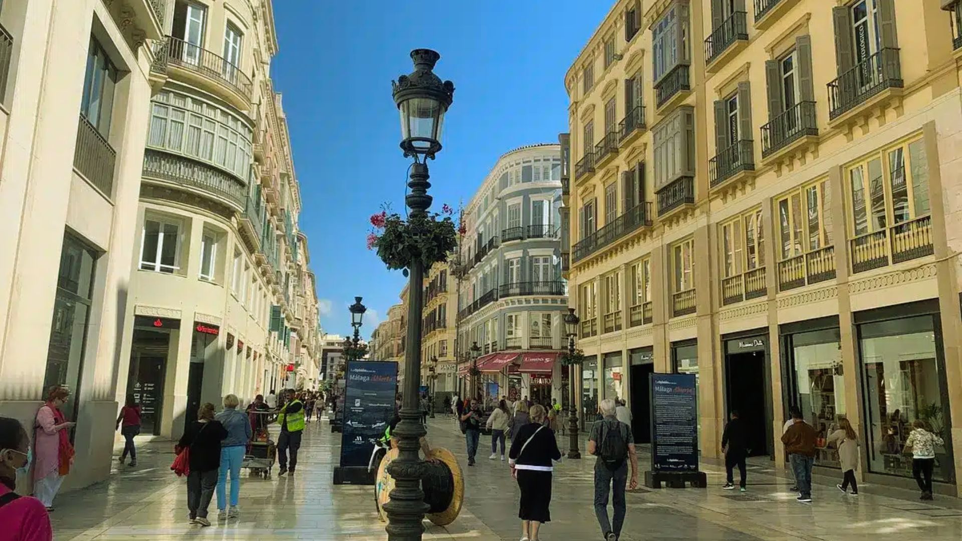 A bustling pedestrian street with historic European buildings, ornate lamp posts, and people walking leisurely under a clear blue sky.