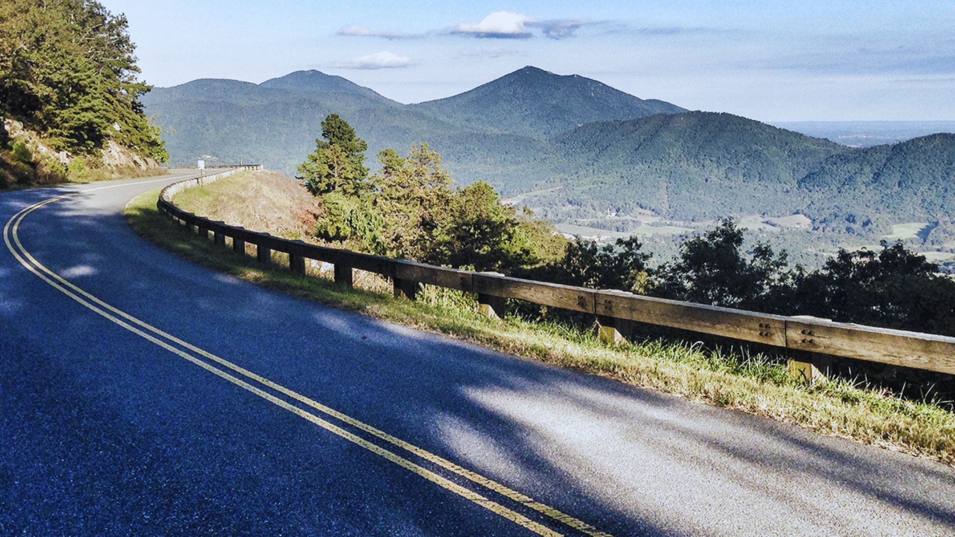 A winding mountain road with yellow lines, flanked by trees and a wooden guardrail, overlooks lush green hills under a clear blue sky. Serene and scenic.