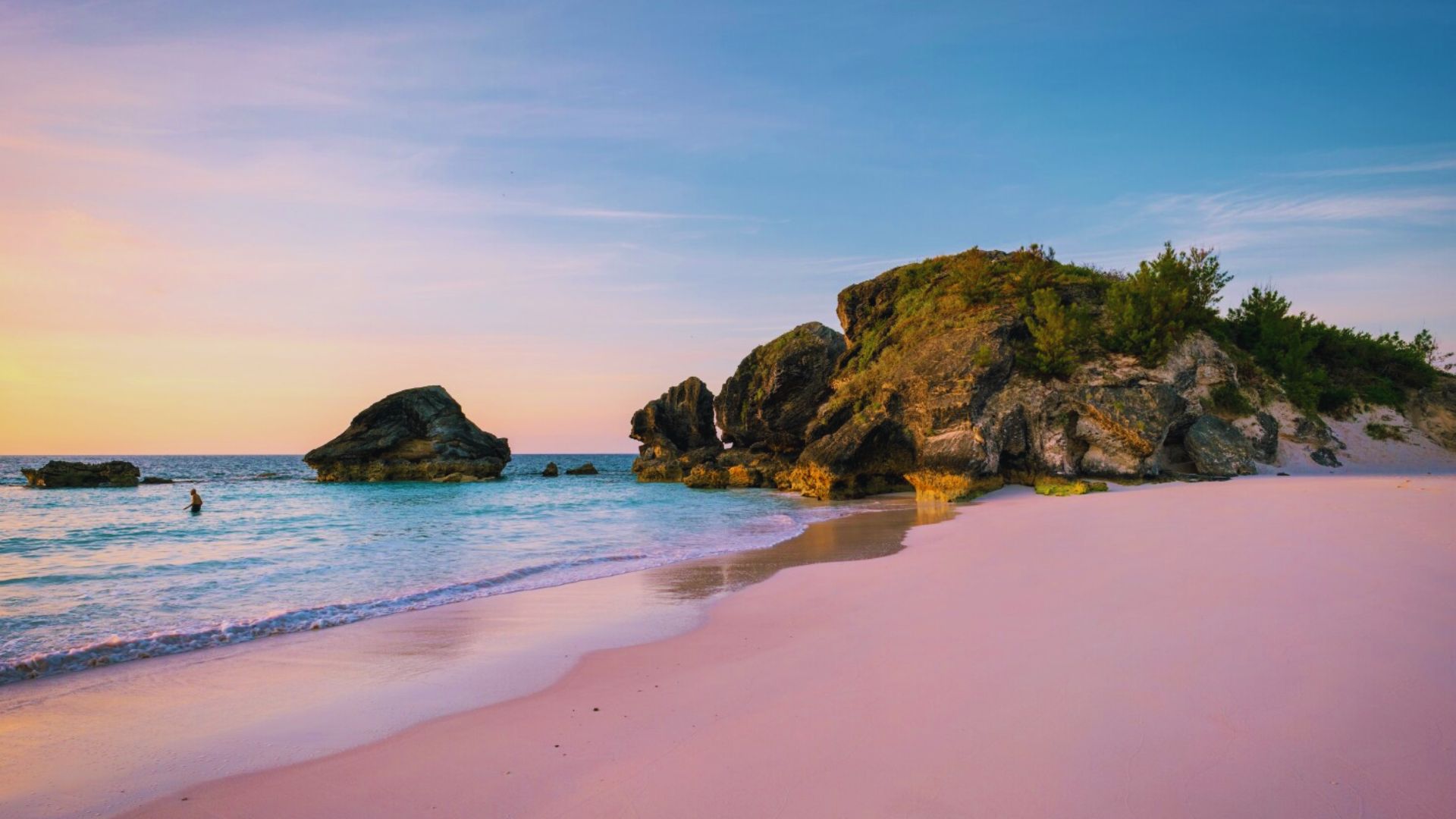 A serene beachscape at sunset with pink sand and gentle waves. Large rock formations sit on the shore, with a swimmer in the turquoise water. Calm, peaceful ambiance.