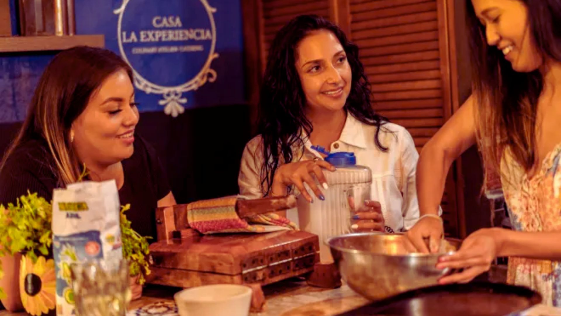 Three women are gathered in a cozy kitchen, smiling and engaging while cooking. One stirs a bowl, creating a warm and collaborative atmosphere.