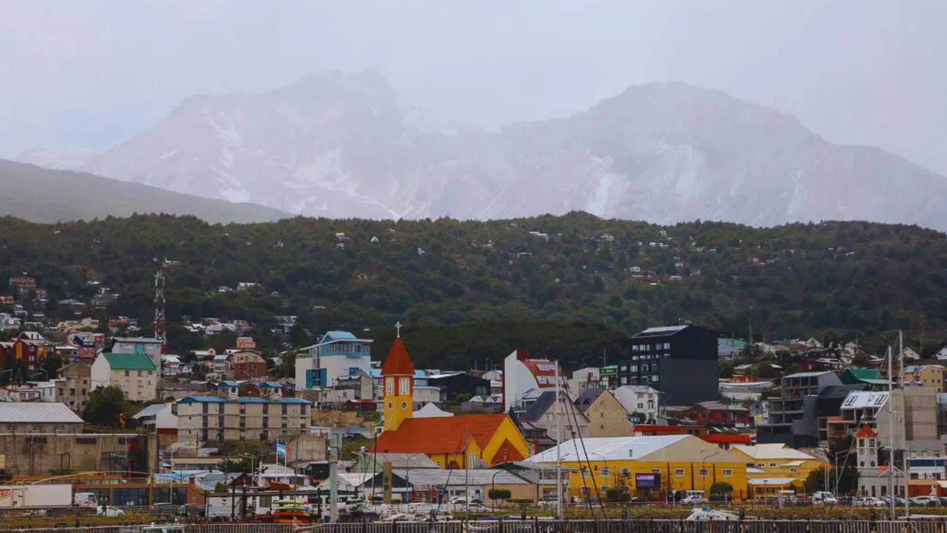 A vibrant coastal town with colorful buildings, including a red-and-yellow church. Misty mountains rise in the background, creating a serene atmosphere.