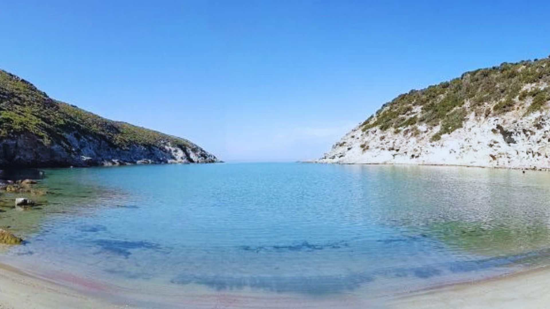 Tranquil bay with clear blue water between two green hills under a bright blue sky, creating a serene and peaceful atmosphere.