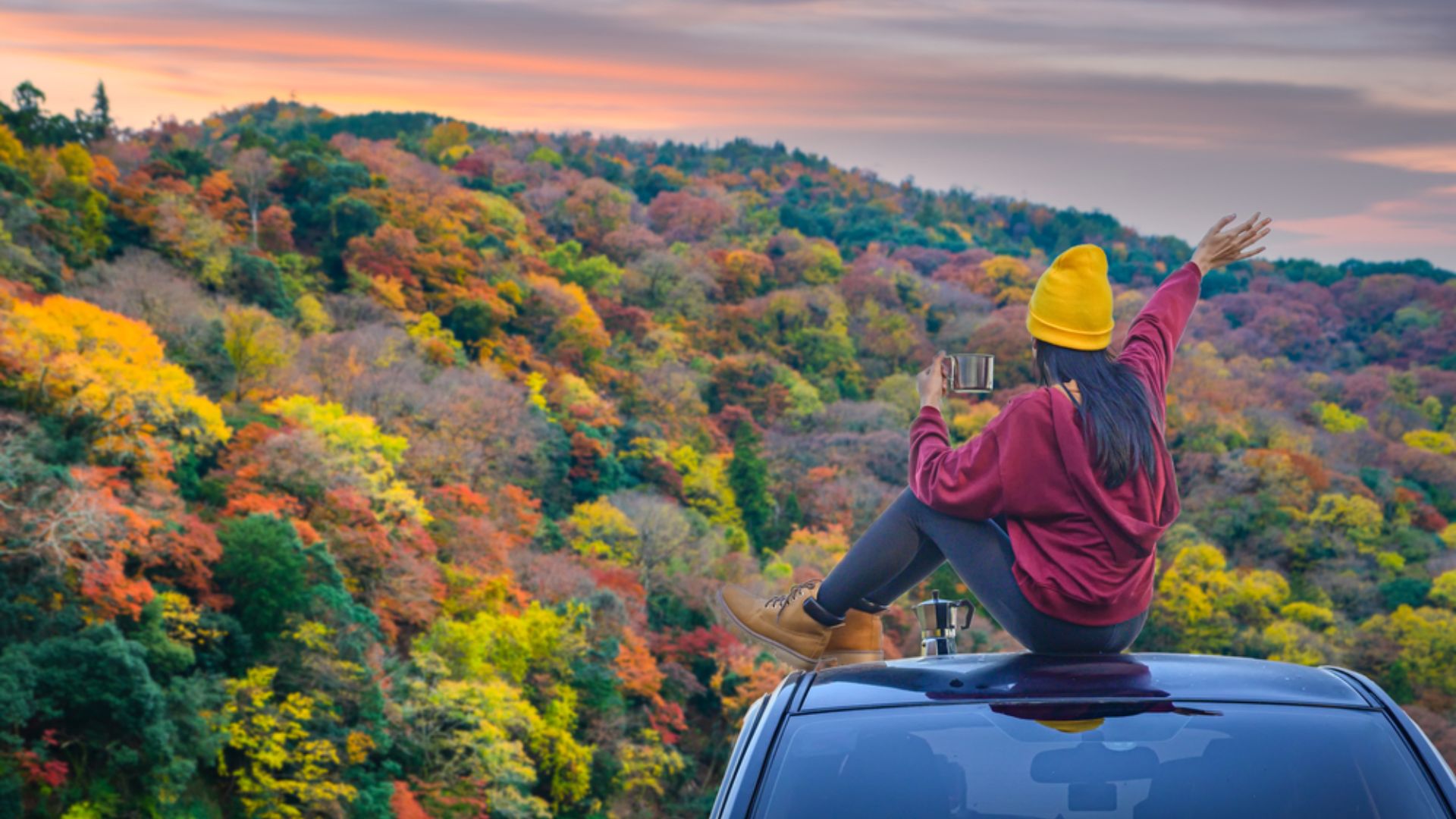 A woman sits on a car with her arms raised, expressing joy and freedom under a clear blue sky.