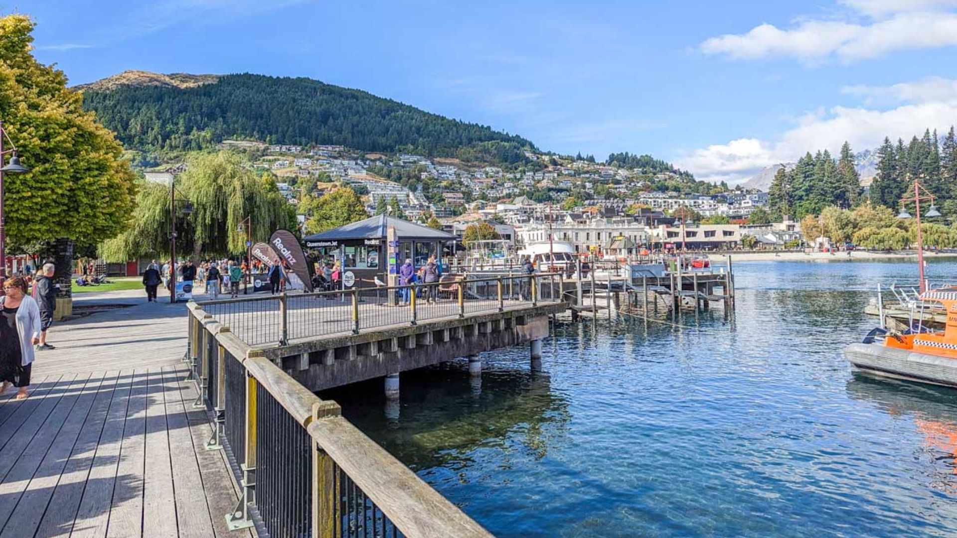 People stroll along the waterfront in Queenstown, New Zealand, with scenic views of the lake and mountains in the background.