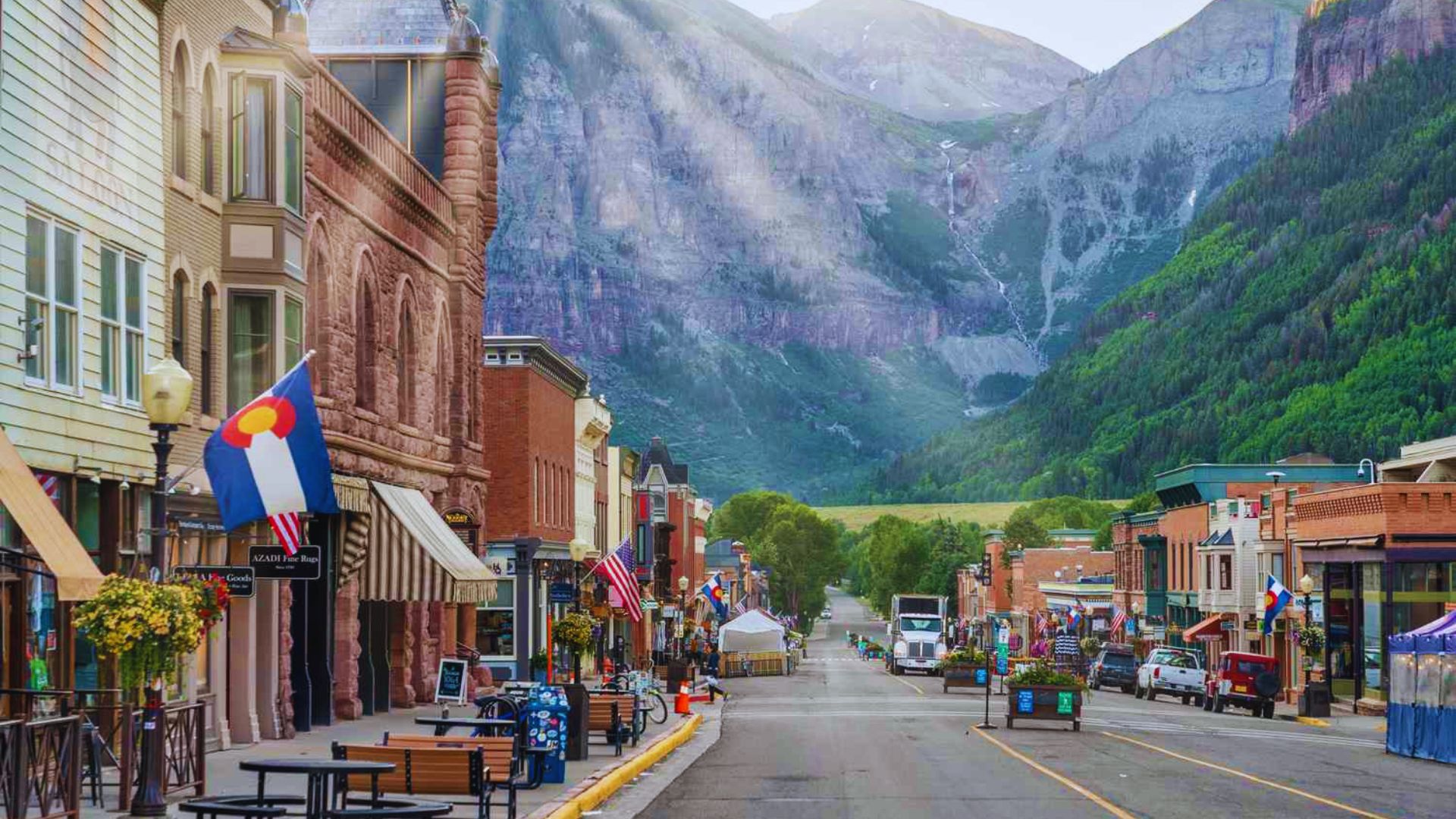 A city street lined with buildings, featuring majestic mountains in the background under a clear blue sky.