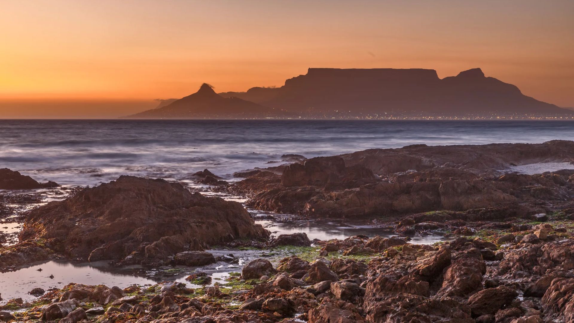 Sunset view of a rocky shoreline with waves, set against a silhouetted mountain range. The sky glows with warm orange hues, creating a tranquil atmosphere.