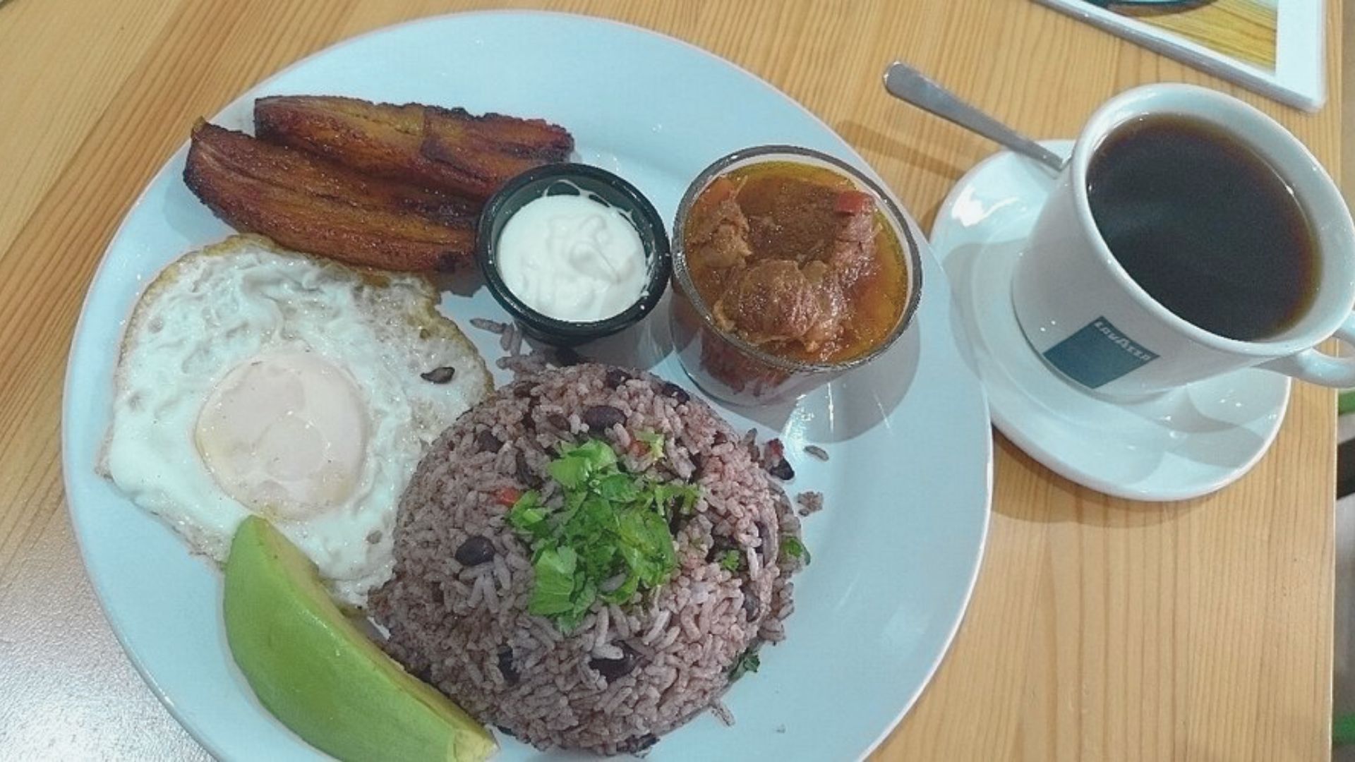 A plate of Costa Rican breakfast featuring gallo pinto, a fried egg, plantains, sour cream, beef stew, avocado, and a cup of black coffee on the side.