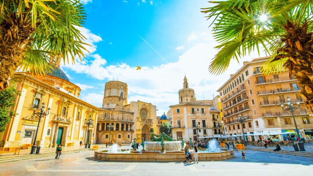 Sunny plaza in Valencia with historic buildings, palm trees, and a large central fountain. The vibrant scene evokes a lively, warm atmosphere.