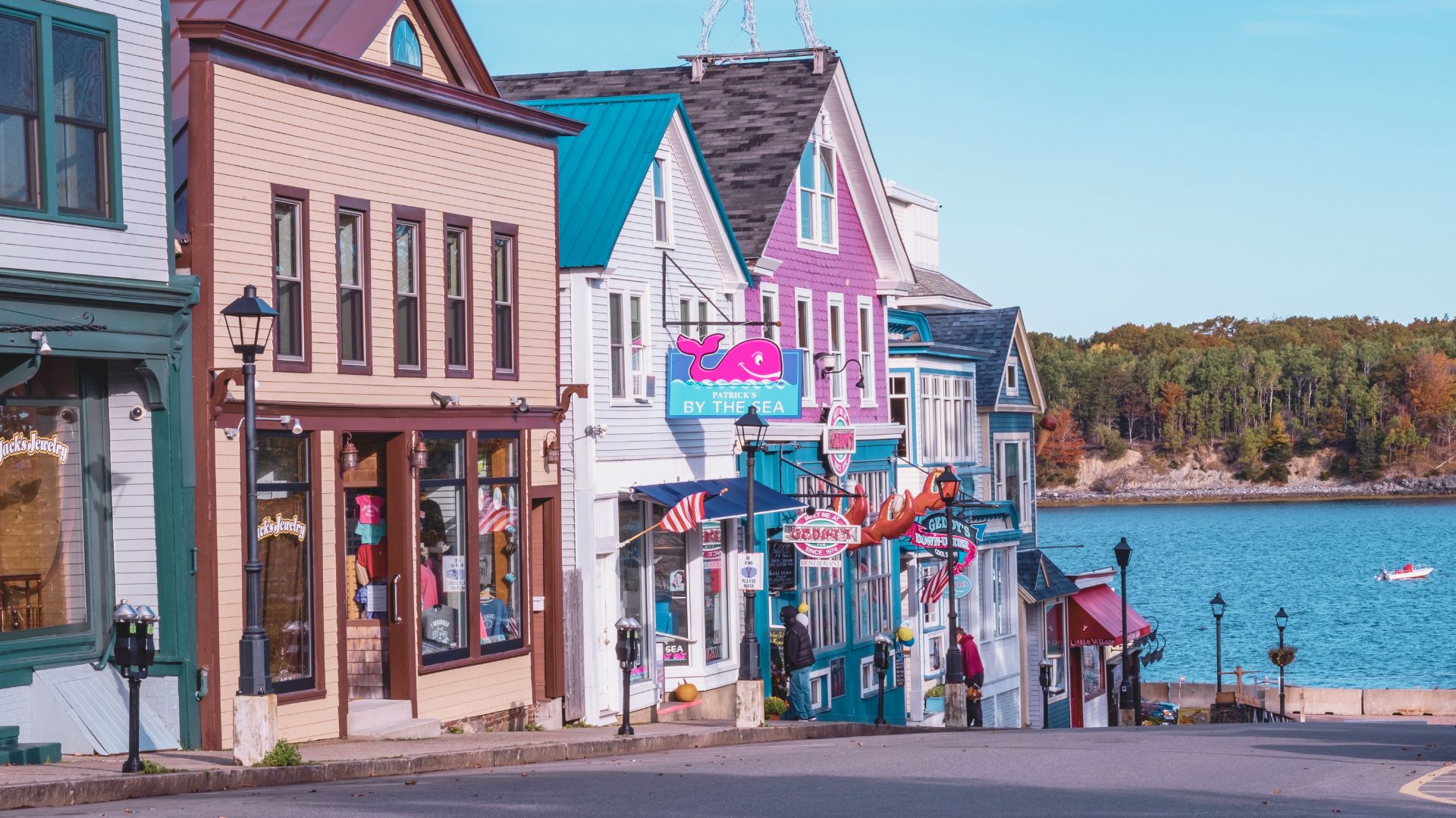 Colorful seaside shops line a sloping street, with vibrant facades and nautical decor. A calm blue bay and forested shoreline are in the background.
