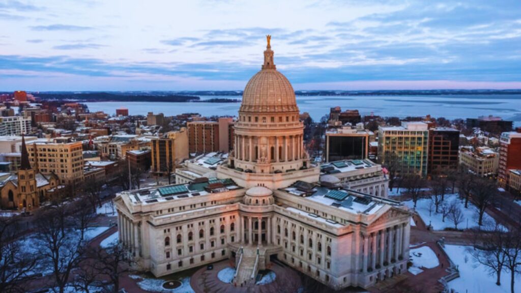 Aerial view of a domed capitol building surrounded by snow-dusted trees and urban buildings at sunset, with a distant lake under a cloudy sky.