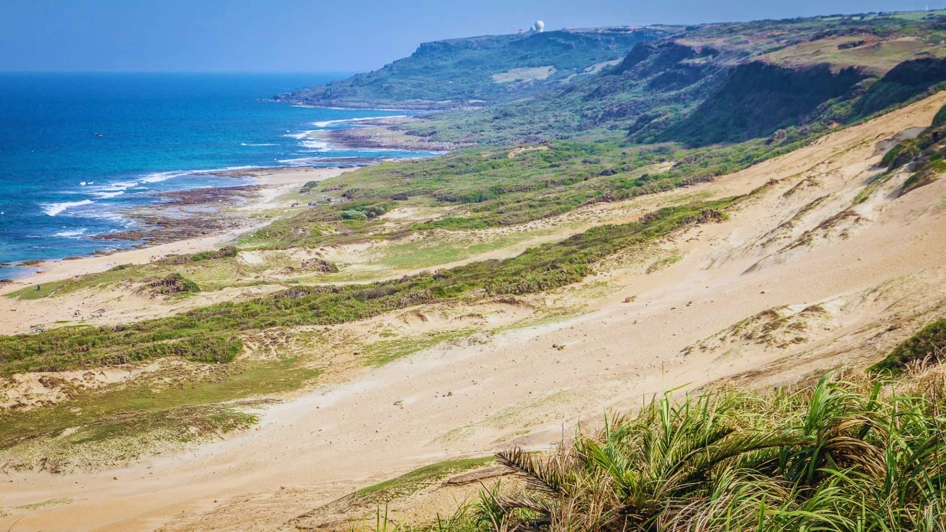 A scenic coastal landscape with sandy dunes and green vegetation, bordered by a deep blue ocean on the left and rugged cliffs in the distance.