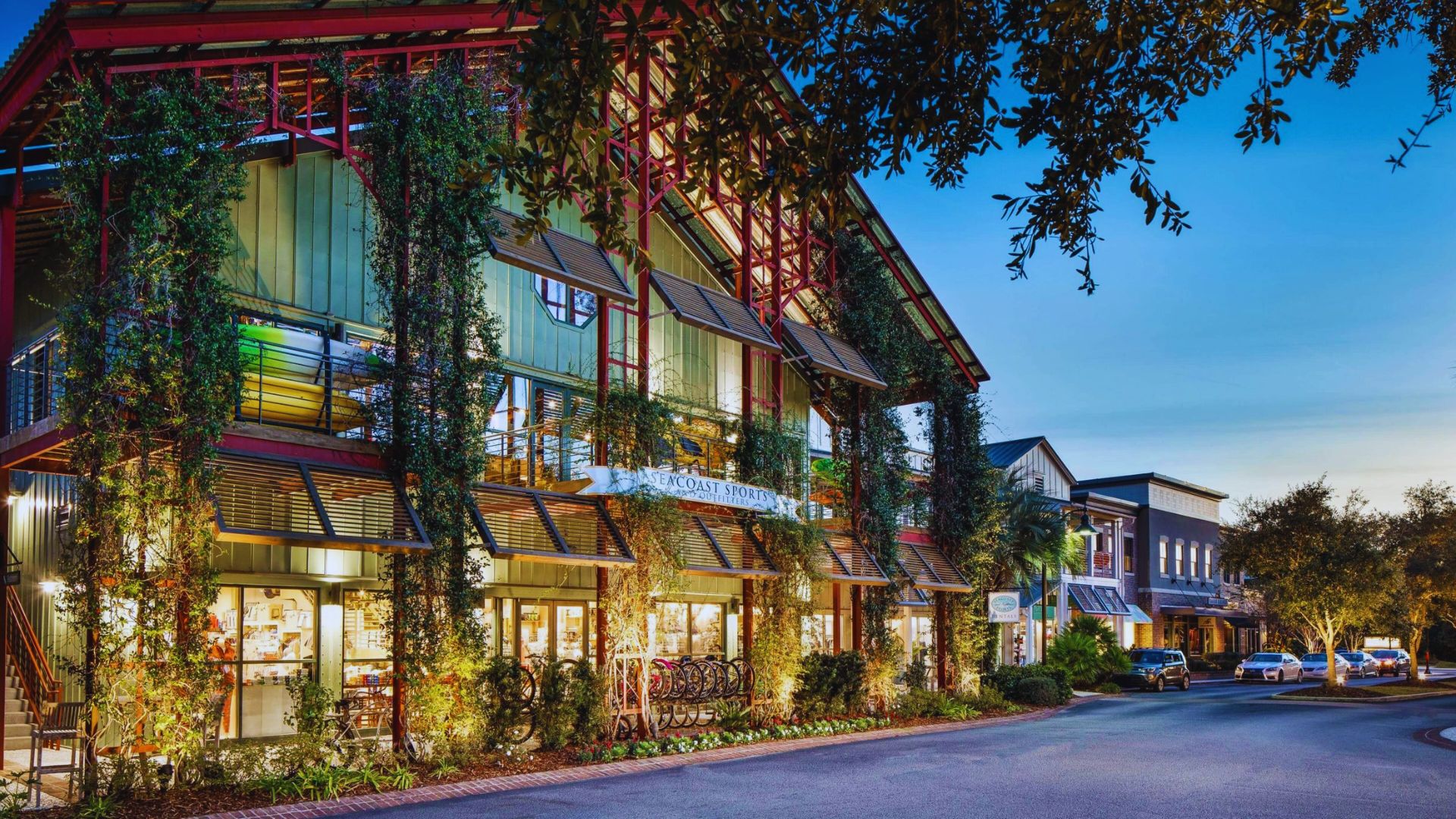 A vibrant street scene at dusk, featuring a two-story building adorned with greenery and large windows illuminated from within. The atmosphere is warm and inviting.