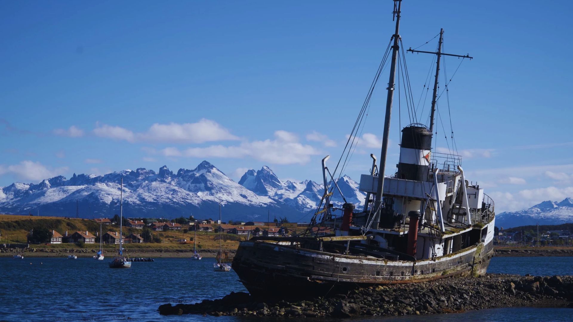 A weathered shipwreck rests on rocky shorelines with a backdrop of snow-capped mountains under a clear blue sky, conveying a serene, timeless mood.