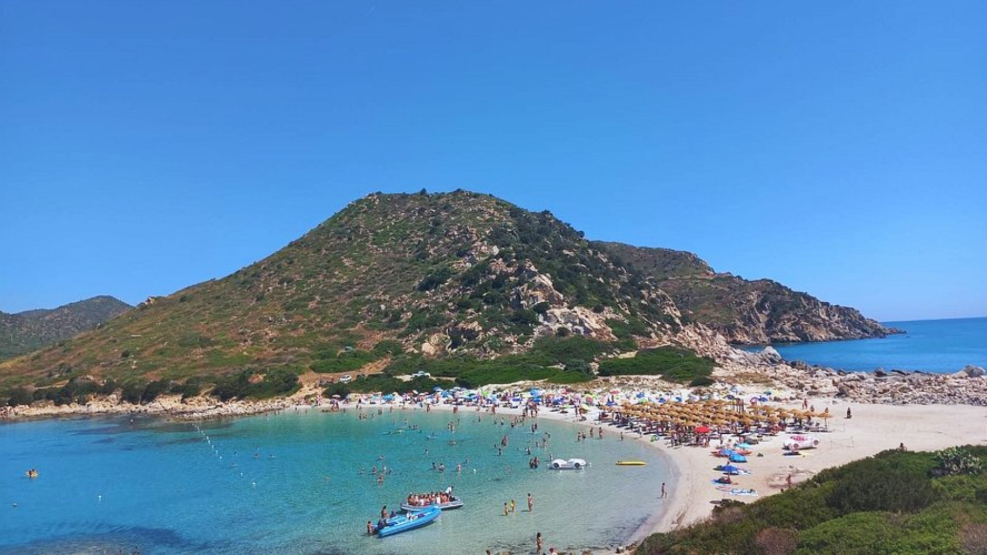 A scenic beach with clear blue water is crowded with people swimming and sunbathing. Green hills and rocky cliffs surround the sandy shore.
