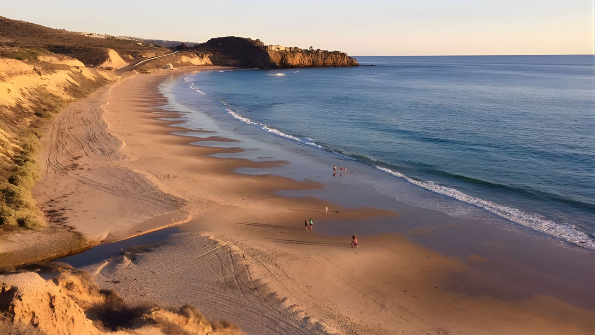 A scenic beach with people walking along the shore, framed by a towering cliff in the background.