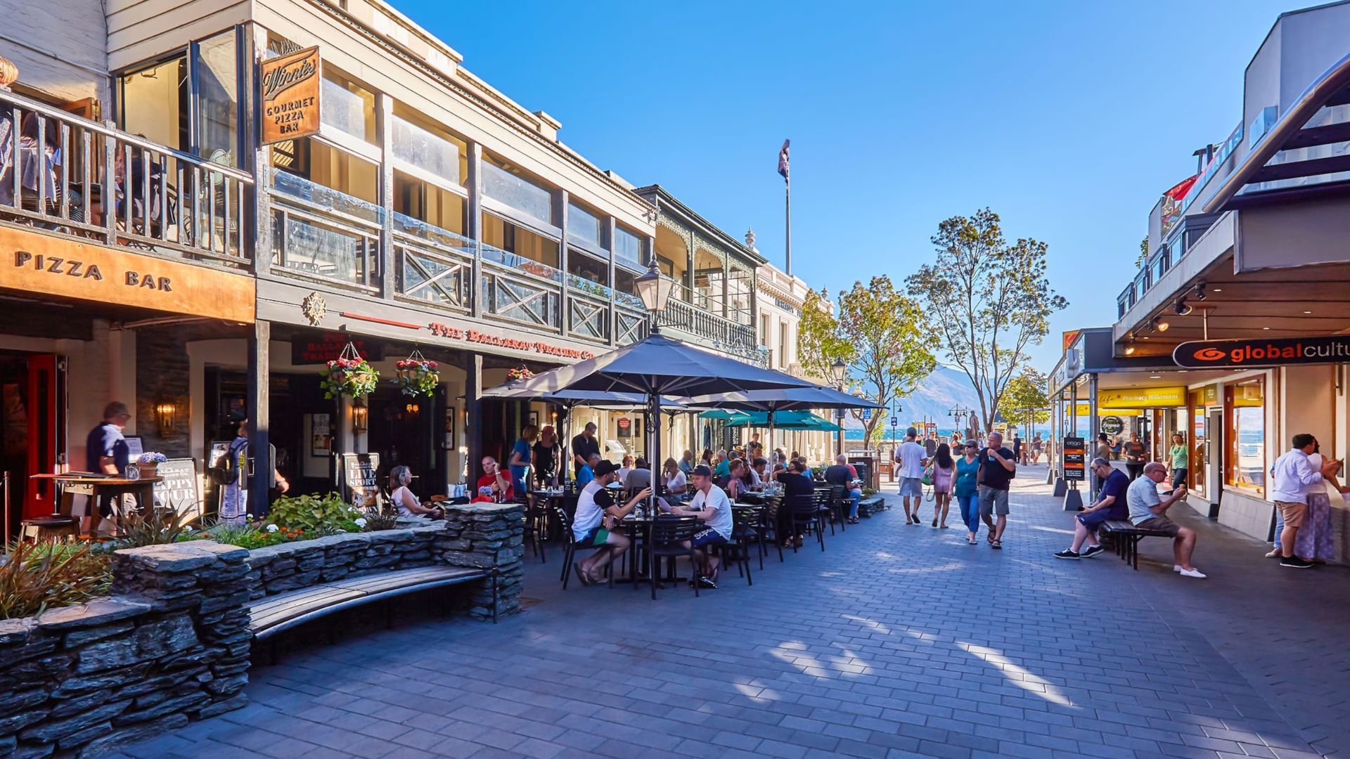 A bustling street scene with pedestrians walking and people relaxing on benches under a clear sky.