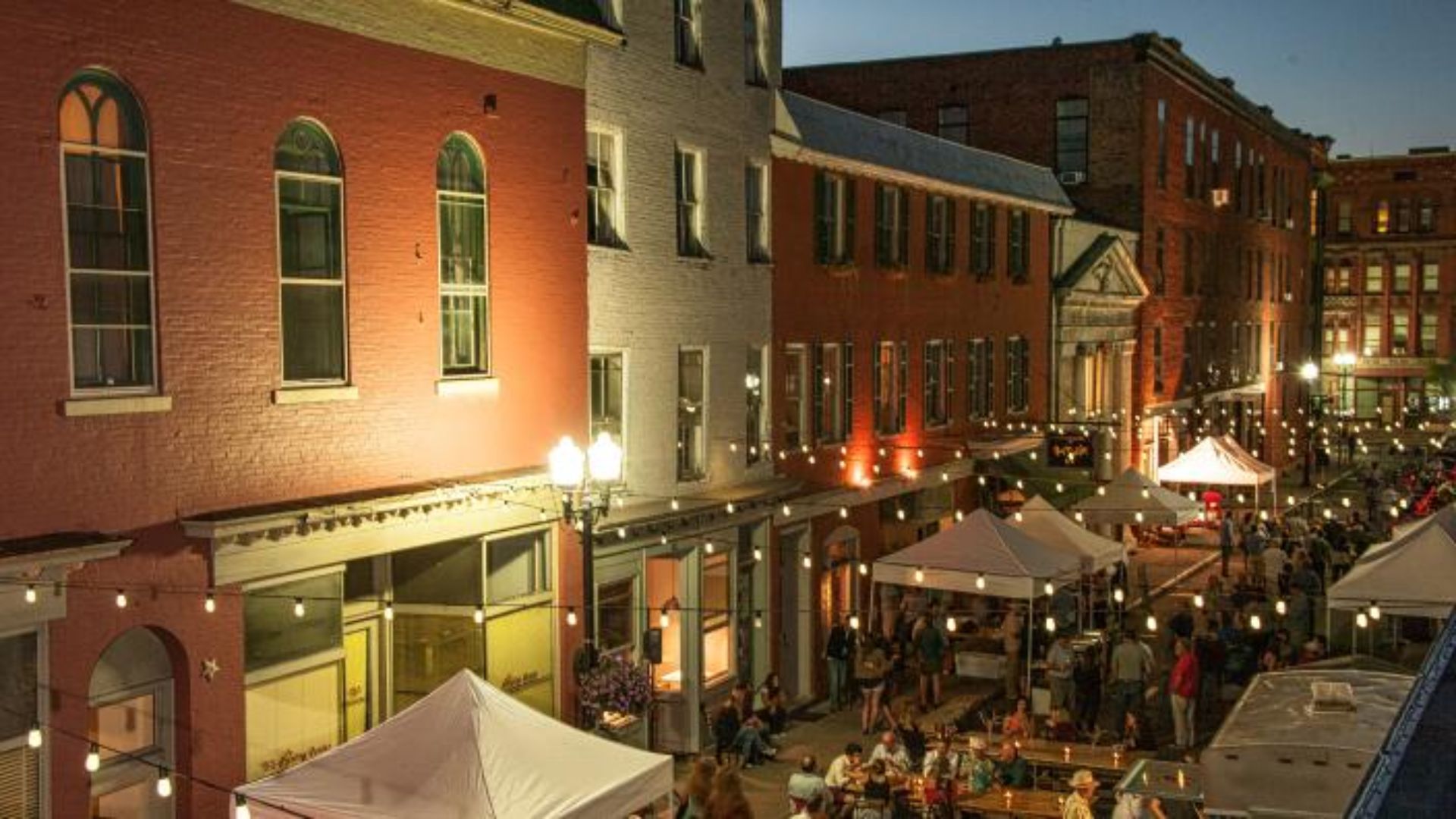 A city street at night featuring tents and tables, illuminated by streetlights and surrounded by urban buildings.