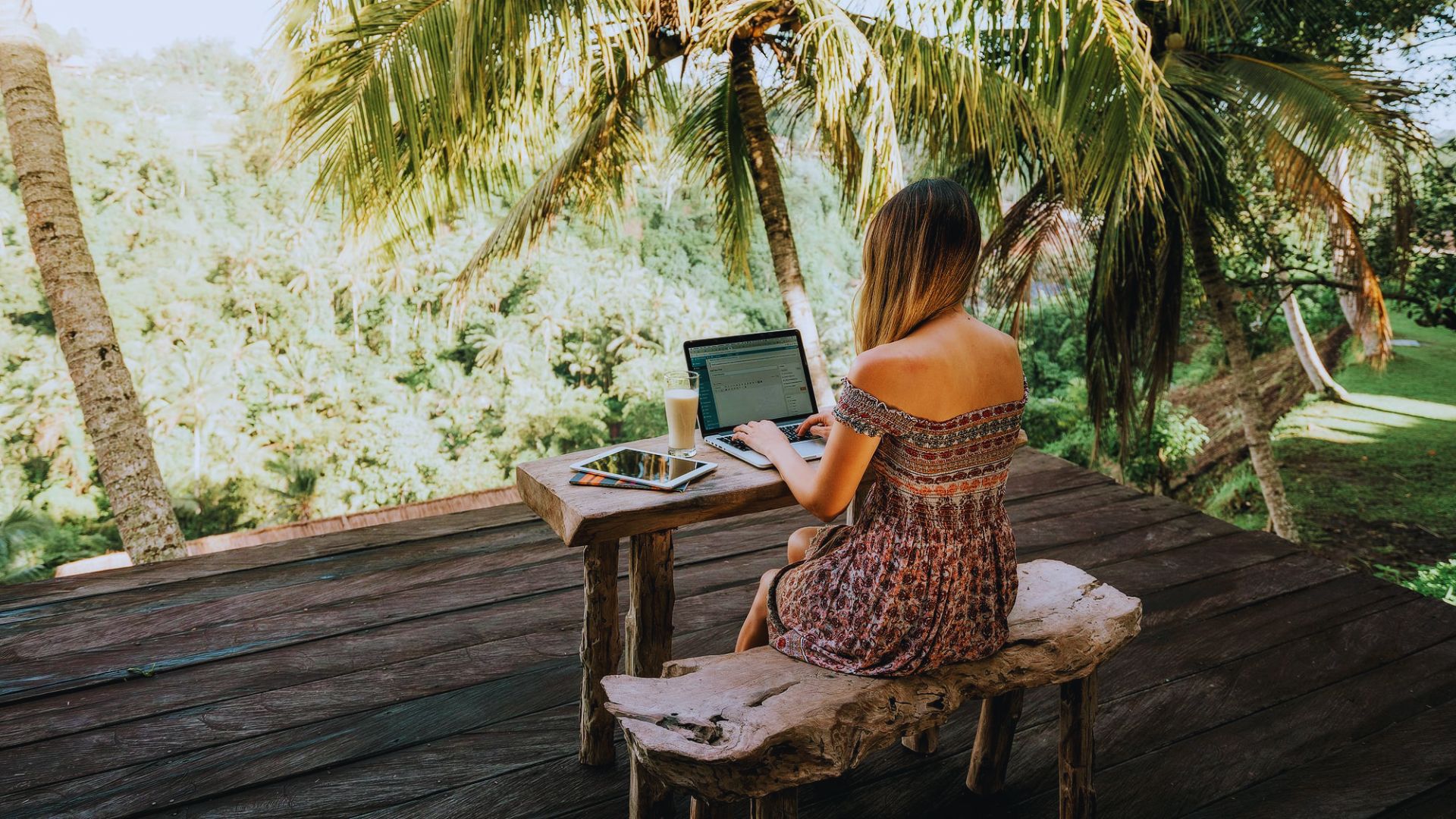 Woman working on a laptop surrounded by tropical plants and a sunny beach in the background.