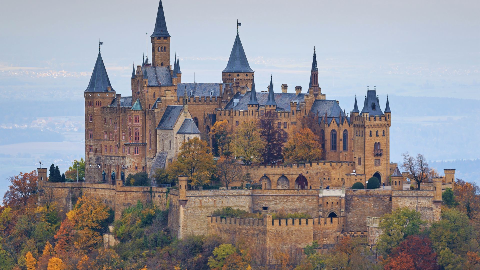 Majestic castle with tall spires atop a hill surrounded by autumn trees, displaying orange and gold foliage. The scene conveys a sense of grandeur and history.